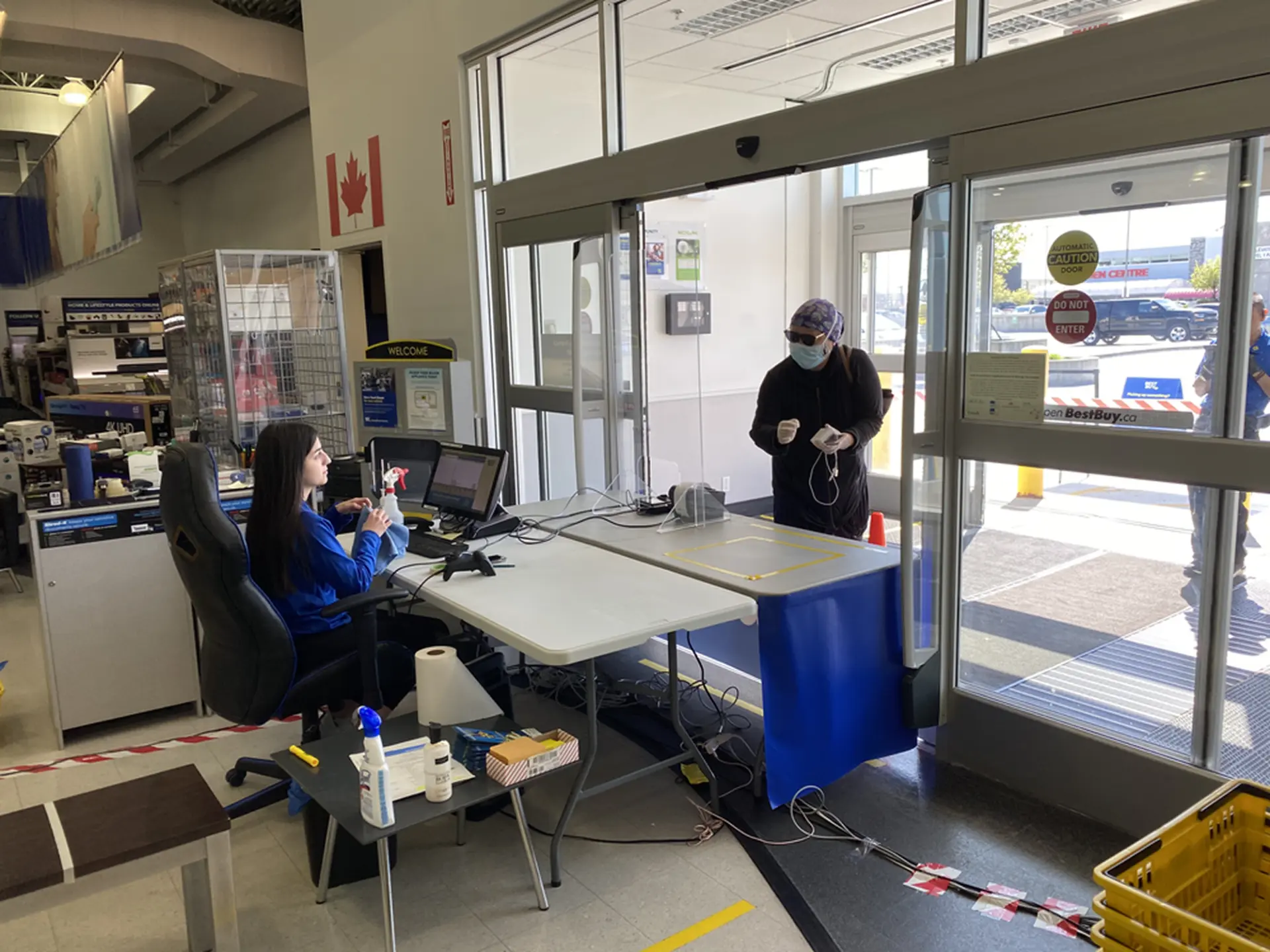 Inside the store entrance, an employee seated at a temporary pickup desk with monitors and hand sanitizer helps a masked customer standing outside behind a clear protective barrier