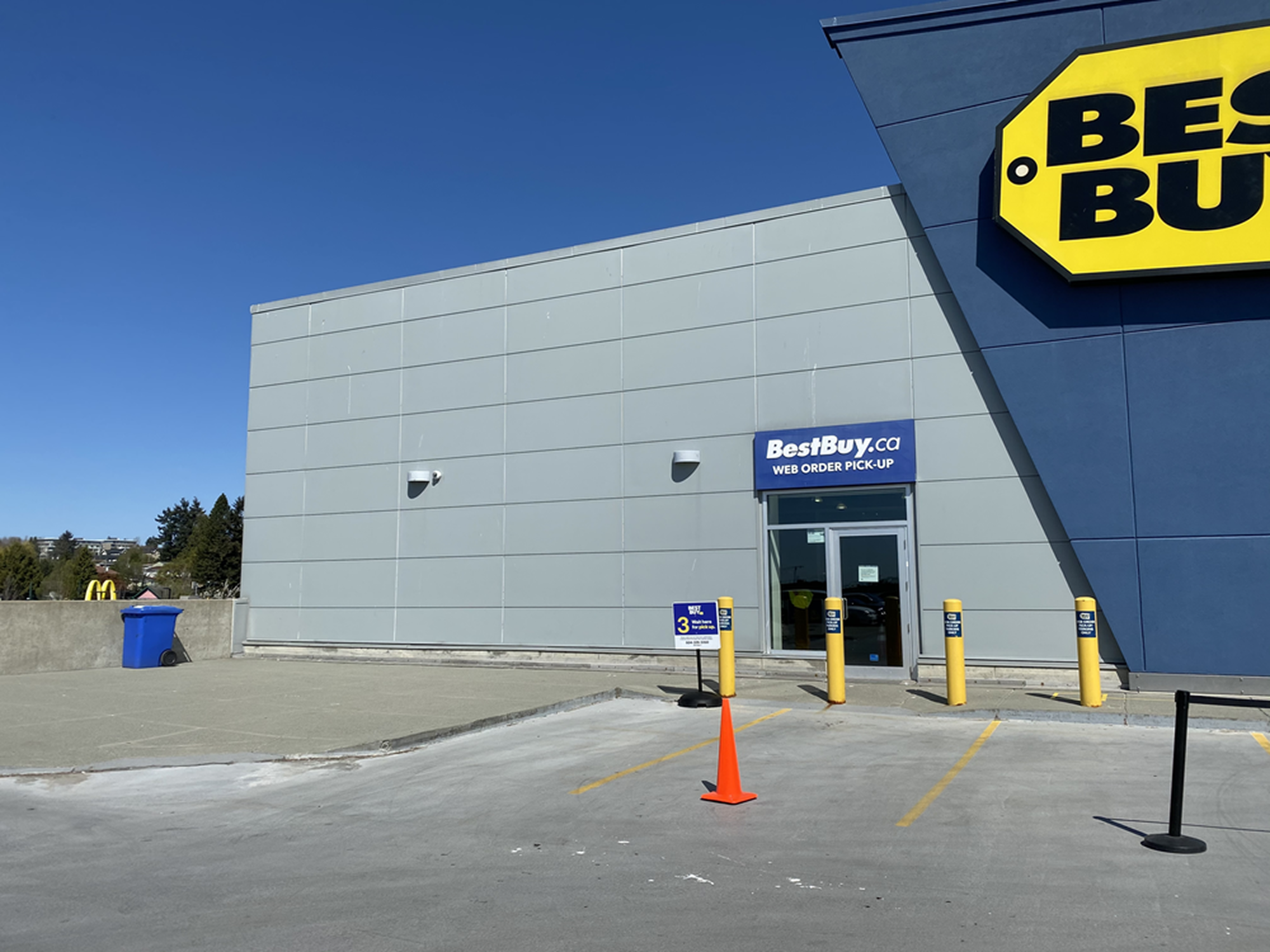 Best Buy building exterior with a blue 'BestBuy.ca Web Order Pick-Up' entrance door, a numbered parking stall marked with an orange cone, and yellow safety bollards