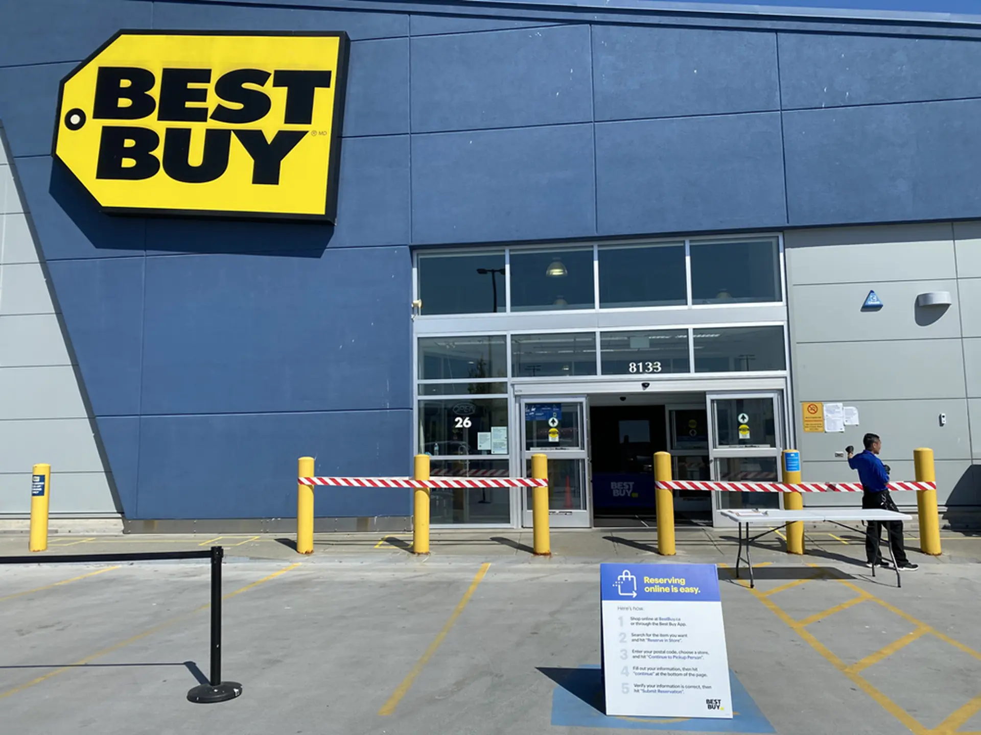 Best Buy store entrance closed off with red-and-white barrier tape between yellow bollards, with a pickup instruction sign, a folding table and an employee standing nearby